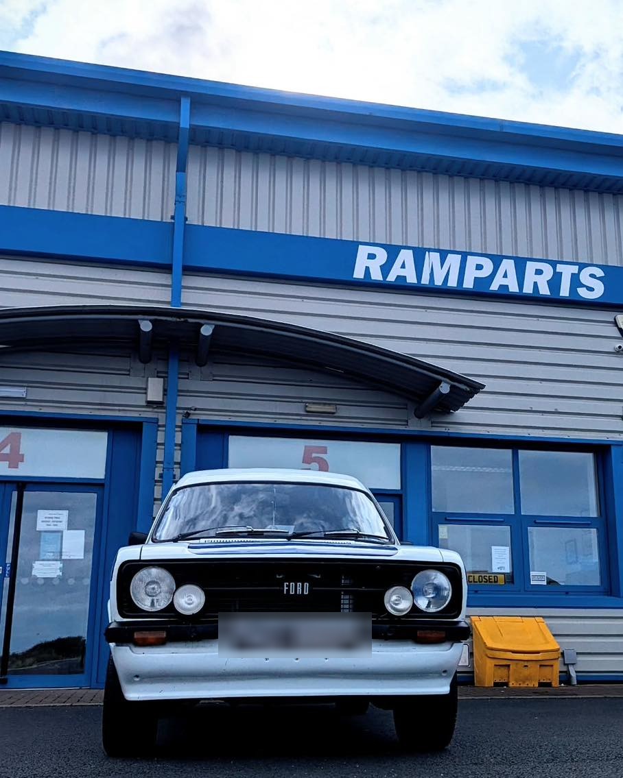 A classic white Ford Escort parked at the Ramparts entrance under the blue RAMPARTS sign